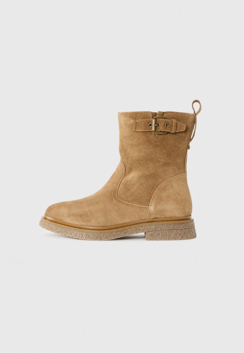 Beige Wildleder-Ankle-Boot mit runder Zehenpartie, strukturiertem Gummisole, seitlichem Schnallen-Detail und Zugschlaufe hinten für einfaches An- und Ausziehen.