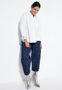 Witte button-up blouse met een pocket en zilveren pailletten manchetten, gecombineerd met donkerblauwe baggy broek versierd met witte kralen en metallic zilveren enkellaarzen.