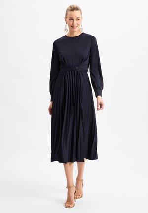 Navy long-sleeve dress with pleated skirt, fitted bodice, and wrap detail at the waist. Smooth fabric, ankle-length, paired with nude heels.