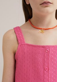 Rozā teksturēta bezpiedurkņu blūze ar viļņainu rakstu un divām rozām pogām, kombinēta ar oranžu pērlīšu kaklarotu ar caurspīdīgu lapas piekariņu.