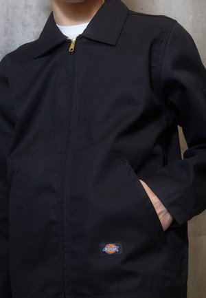 Persona che indossa una giacca nera con zip e colletto, mano in tasca e logo visibile del marchio Dickies vicino all'orlo inferiore.