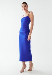 Modrá bodycon šaty s tenkými ramínky, nařasenými detaily a midi délkou. Nosené s zlatými sandály na vysokém podpatku. Jednoduchá textura.