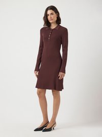 Femme debout portant une robe en tricot marron ajustée à manches longues avec des boutons et des escarpins noirs à bride arrière sur un fond blanc.