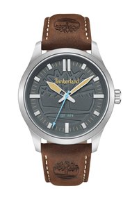 Timberland laikrodis su nerūdijančio plieno korpusu, pilku tekstūruotu ciferblatu, sidabro spalvos valandų žymekliais ir rudu odiniu dirželiu su įspaustu logotipu. Mėlynas sekundinis rodyklė.