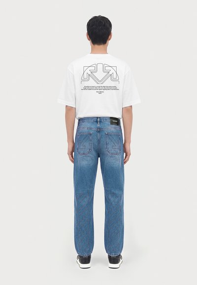 Un homme est debout de dos, portant un t-shirt blanc à motif graphique, un jean bleu avec des dessins au dos et sur les jambes, et des baskets noires et blanches sur un fond uni.
