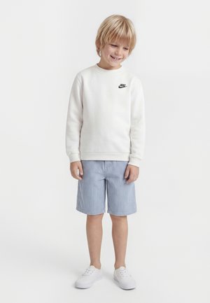 Lächelnder Junge trägt weißen Nike-Sweatshirt, blau gestreifte Shorts und weiße Turnschuhe, steht vor einem schlichten hellen Hintergrund.