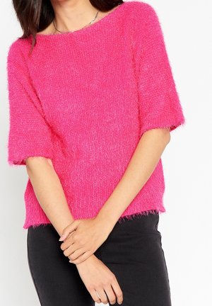 Kobieta ubrana w jasnoróżowy, puszysty sweter z krótkim rękawem i czarne spodnie, z założonymi na wysokości talii skrzyżowanymi ramionami, na jednolitym białym tle.