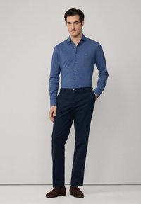 Blaues Hemd mit langen Ärmeln, marineblaue Hose und braune Wildlederschuhe. Das Hemd hat eine dezente Textur und ein kleines Logo auf der Brust.