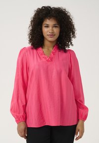 Blouse rose à manches longues avec un décolleté en V froncé, tissu texturé créant des rayures verticales, et poignets élastiques. Un pantalon noir est porté en dessous.