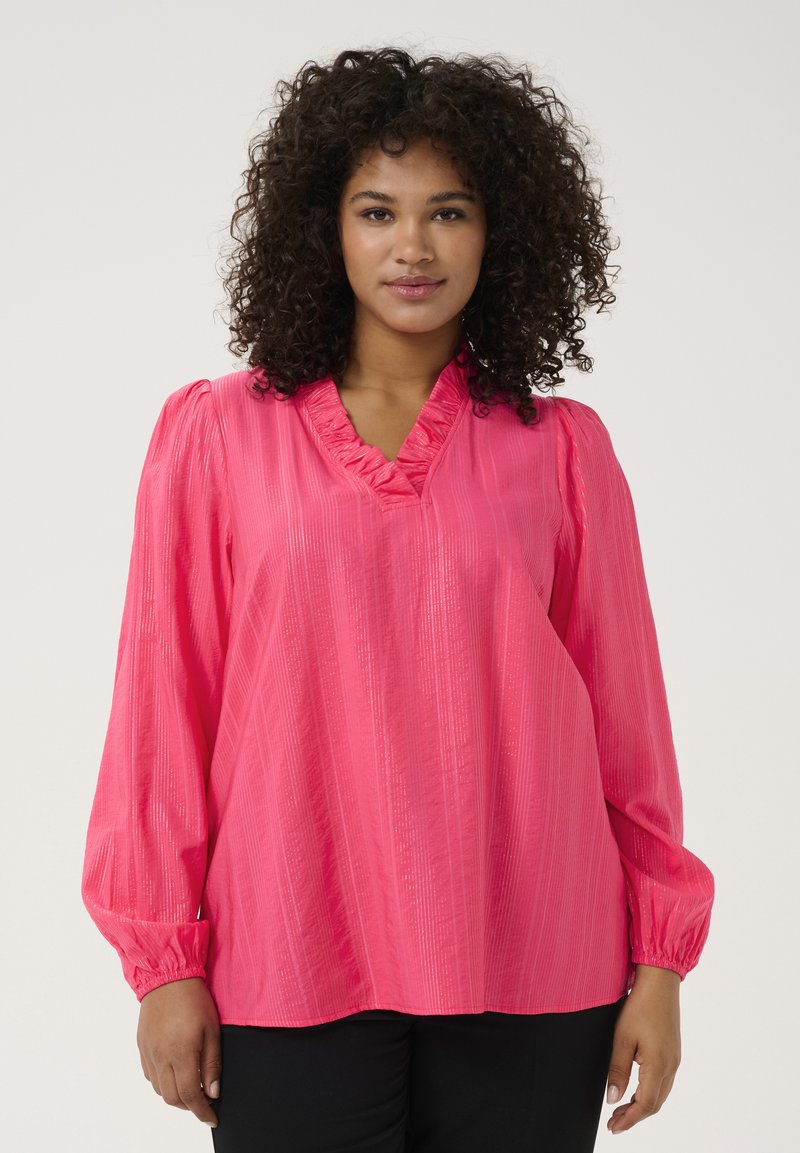 Blouse rose à manches longues avec un décolleté en V froncé, tissu texturé créant des rayures verticales, et poignets élastiques. Un pantalon noir est porté en dessous.
