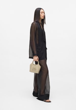 Femme portant un long blazer noir transparent et un pantalon, tenant un petit sac à main beige, debout de profil sur fond blanc.