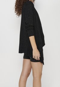 Svart cardigan med lös passform, dekorerad med flerfärgade prickar, ribbade ärmar och asymmetrisk fåll, bärs över svarta shorts.