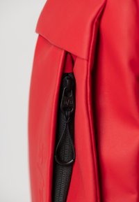 Zaino rosso con una finitura liscia e opaca, dotato di una zip nera prominente e dettagli in corda laterali per una chiusura sicura.