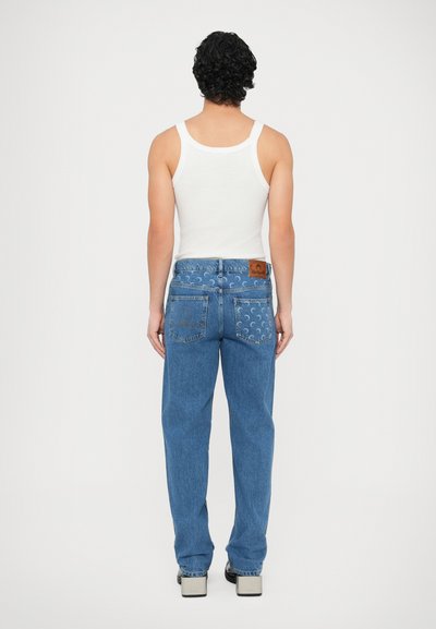 Modre jeans hlače iz denima z rahlim krojem, vidnimi šivi, zadnjimi žepi in rjavim usnjenim vložkom na pasu. Nošene z belo majico brez rokavov.