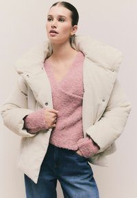 Weißes Corduroy-Puffermantel mit großem Kragen, Druckknopfverschluss und rosa strukturiertem Pullover darunter; kombiniert mit blauen Jeans.