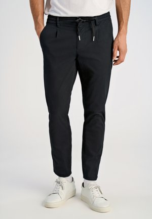 Mand iført sorte, tætsiddende bukser med snøring i taljen, hvide sokker og hvide sneakers, stående mod en ensformig lys baggrund.