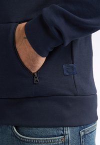 Navy blue sweatshirt with a front pocket featuring a zipper, ribbed cuffs, and a small branded label on the lower side.
