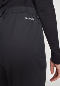 Nahaufnahme einer Person, die schwarze Hosen mit elastischem Bund trägt und das Wort TEXAPORE nahe der Taille aufgedruckt hat.