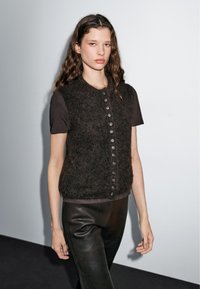 Frau mit langen lockigen Haaren, die eine schwarze, flauschige, geknöpfte Weste über einem kurzärmeligen schwarzen Hemd und einer schwarzen Lederhose trägt, vor einem einfarbigen Hintergrund.