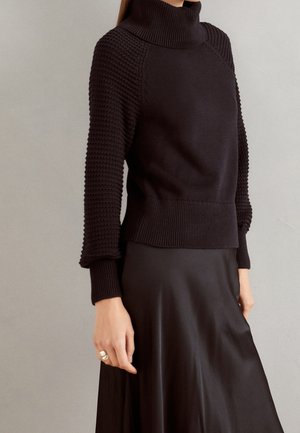 Sort tekstureret strikket rullekravesweater med ribkanter kombineret med en sort satin nederdel, båret af en person mod en neutral baggrund.