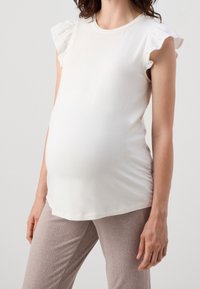 Blusa de maternidad blanca con mangas cortas con encaje, material de algodón suave. Combinada con pantalones beige de canalé ligeros. Ajuste simple y relajado.