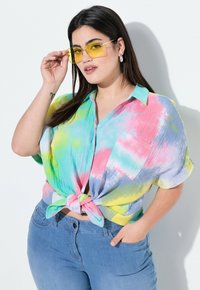 Maglietta tie-dye multicolore con maniche corte, che presenta tonalità di rosa, blu, verde e giallo, annodata in vita. Abbinata a jeans azzurri.