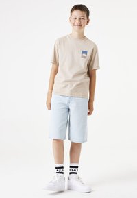 Beige Baumwoll-T-Shirt mit einem bunten Patch, hellblaue Denim-Shorts, weiße Sneaker und schwarz-weiß gestreifte Socken.