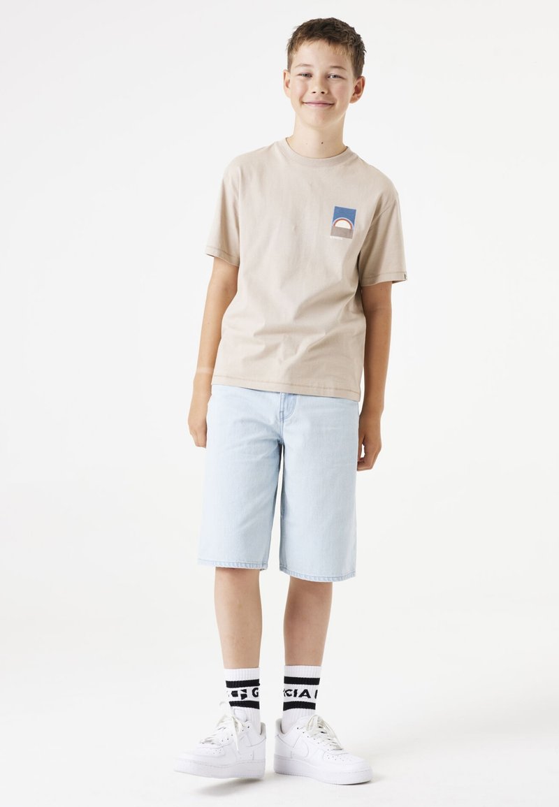 Beige Baumwoll-T-Shirt mit einem bunten Patch, hellblaue Denim-Shorts, weiße Sneaker und schwarz-weiß gestreifte Socken.