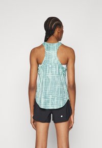 Kvinne med flettet hår iført en blåmønstret racerback tanktopp og svarte treningsshorts, stående med ryggen til mot en ensfarget bakgrunn.