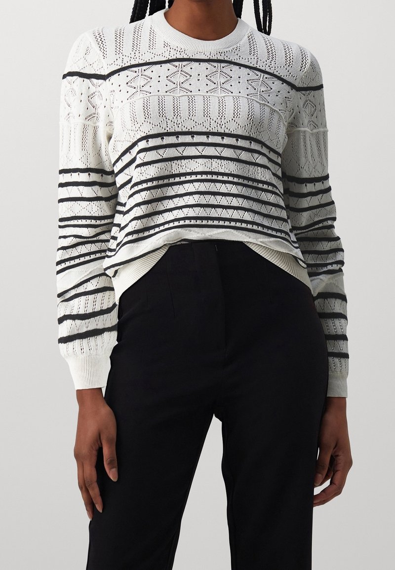 Pull blanc en maille avec des rayures horizontales noires et des motifs géométriques, associé à un pantalon noir taille haute, porté par une personne debout.
