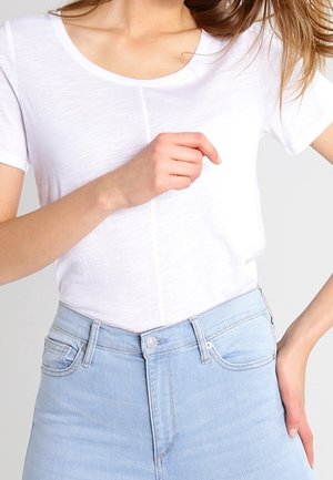 Kobieta ubrana w prostą, białą, krótki rękaw koszulkę włożoną do jasnoniebieskich, wysokich dżinsów, z jedną ręką spoczywającą na biodrze.