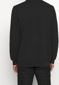 Sudadera negra de manga larga, cuello redondo, con puños y dobladillo de canalé. Textura suave, diseño minimalista, vista trasera lisa. Combinada con pantalones de cuadros.