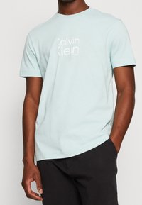 Camiseta de algodón azul claro con un logo blanco de Calvin Klein en el pecho. Manga corta, cuello redondo, textura suave y corte relajado.