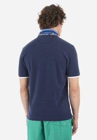 Mörkblå pikétröja med texturerad tyg, försedd med en blå krage med vit text och vita detaljer på ärmkanterna.