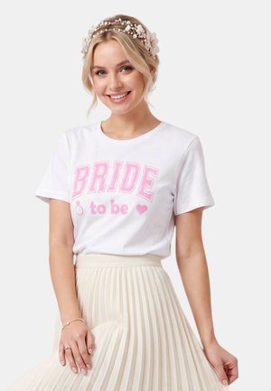 Mujer sonriente con una camiseta blanca "Bride to be", una diadema de perlas y flores, y una falda plisada beige.