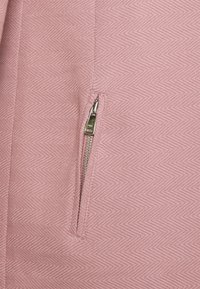 Närmre bild av en delvis dragkedja av metall på strukturerad rosa tyg med ett subtilt diagonalt mönster.