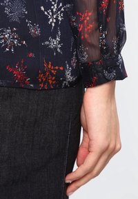 Blusa trasparente a motivi floreali con base blu scuro, caratterizzata da dettagli rossi e bianchi e polsini con bottoni. Abbinata a un capo scuro uniforme.