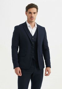 Costume trois pièces marine foncé avec une veste ajustée, des revers crantés, un gilet boutonné et un pantalon sur mesure. Chemise habillée blanche en dessous.