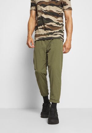 Pánské maskáčové tričko v béžové a zelené barvě, kombinované se zelenými zúženými kalhotami s elastickými manžetami a černými teniskami.