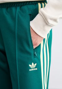 Smaragdfärgade träningsbyxor med gula ränder, utrustade med en ficka med dragkedja och en broderad Adidas-logga på vänster ben. Slät textur.