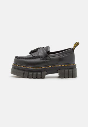 Mocassin en cuir noir avec semelle crantée épaisse, surpiqûres jaunes, détail pompon et franges, et languette de tirage de talon marquée.