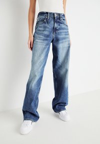 Osoba v volných vybledlých modrých džínách s vysokým pasem a bílými teniskami na jednobarevném bílém pozadí.