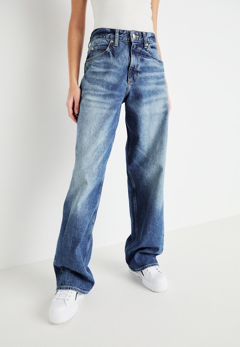 Osoba v volných vybledlých modrých džínách s vysokým pasem a bílými teniskami na jednobarevném bílém pozadí.