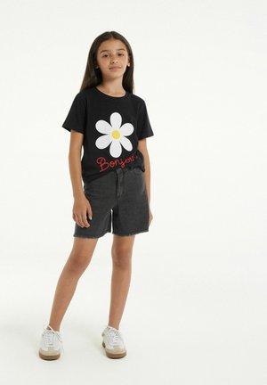 T-shirt noir avec une grande marguerite blanche au centre jaune et le texte rouge "Bonjour", associé à un short en denim noir et des baskets blanches.
