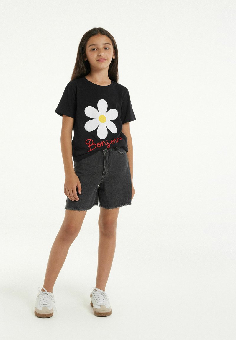 Černé tričko s velkým bílým sedmikráskou se žlutým středem a červeným textem "Bonjour", kombinované s černými džínovými šortkami a bílými teniskami.