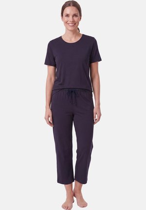 Femme souriante debout pieds nus, portant un t-shirt violet foncé à manches courtes et un pantalon court assorti à la taille avec un cordon de serrage.