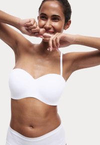 Witte strapless bh met een gladde, gevormde ontwerp en een subtiele V-vormige halslijn. De armen van het model zijn omhoog geheven, waardoor de pasvorm van het kledingstuk zichtbaar is.