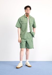 Conjunto de dos piezas verde hecho de una tela ligera, que incluye una camisa de botones de manga corta con bolsillos y pantalones cortos cargo a juego.