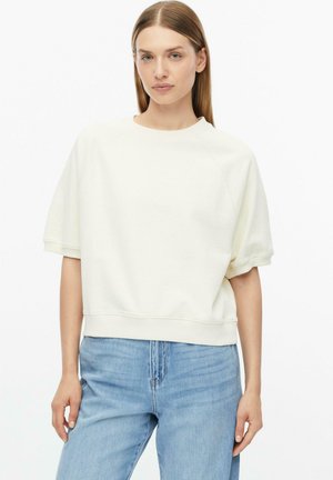 Femme aux cheveux châtain clair et raides porte un haut blanc ample à manches courtes et un jean taille haute bleu clair, debout devant un fond blanc.