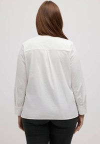 Camicia bianca a maniche lunghe con colletto a punta, dettaglio con piega sul retro e polsini abbottonati, abbinata a jeans scuri. Tessuto liscio e senza pieghe.